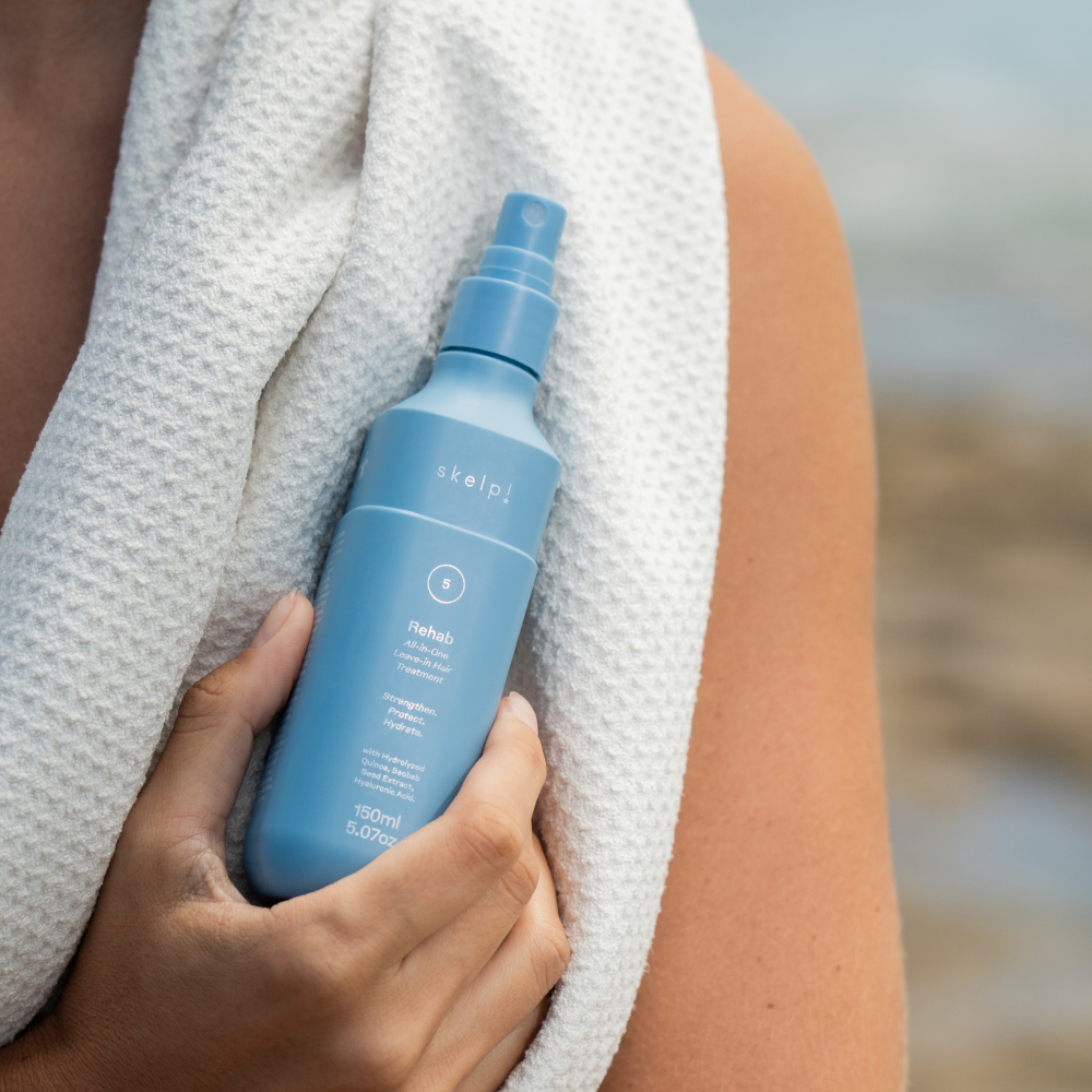 Person holding a skelp! rehab all in one leave in hair treatment against microfibre towel.