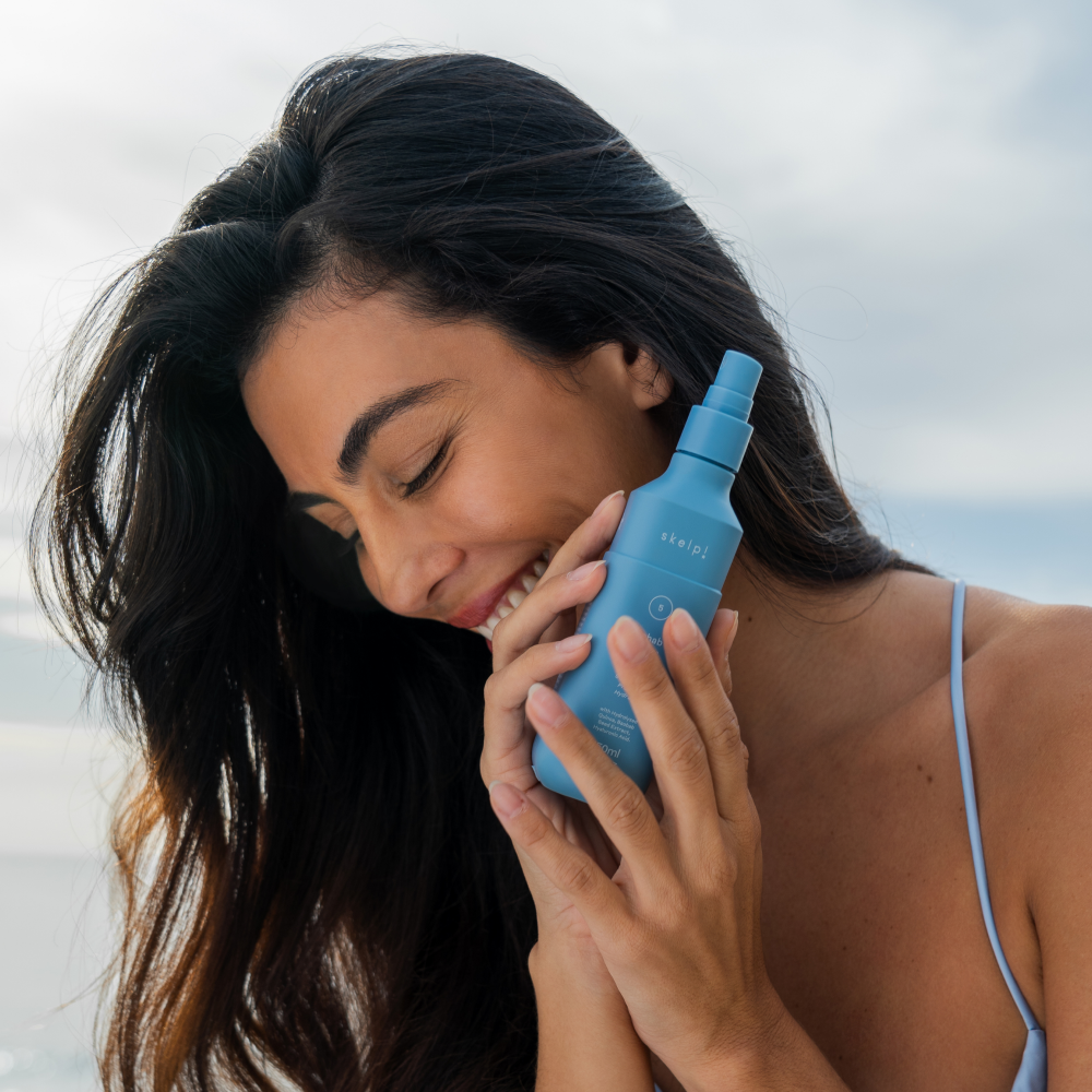 Woman with beautiful brown hair holding a skelp rehab haircare all in one leave in treatment bottle against her face with blue ocean behind.